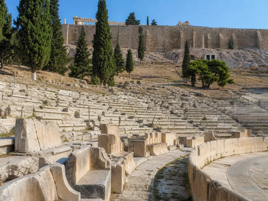 Πέτρινα καθίσματα του Θεάτρου του Διονύσου στη νότια κλιτύ, με τα τείχη της Ακρόπολης από πάνω